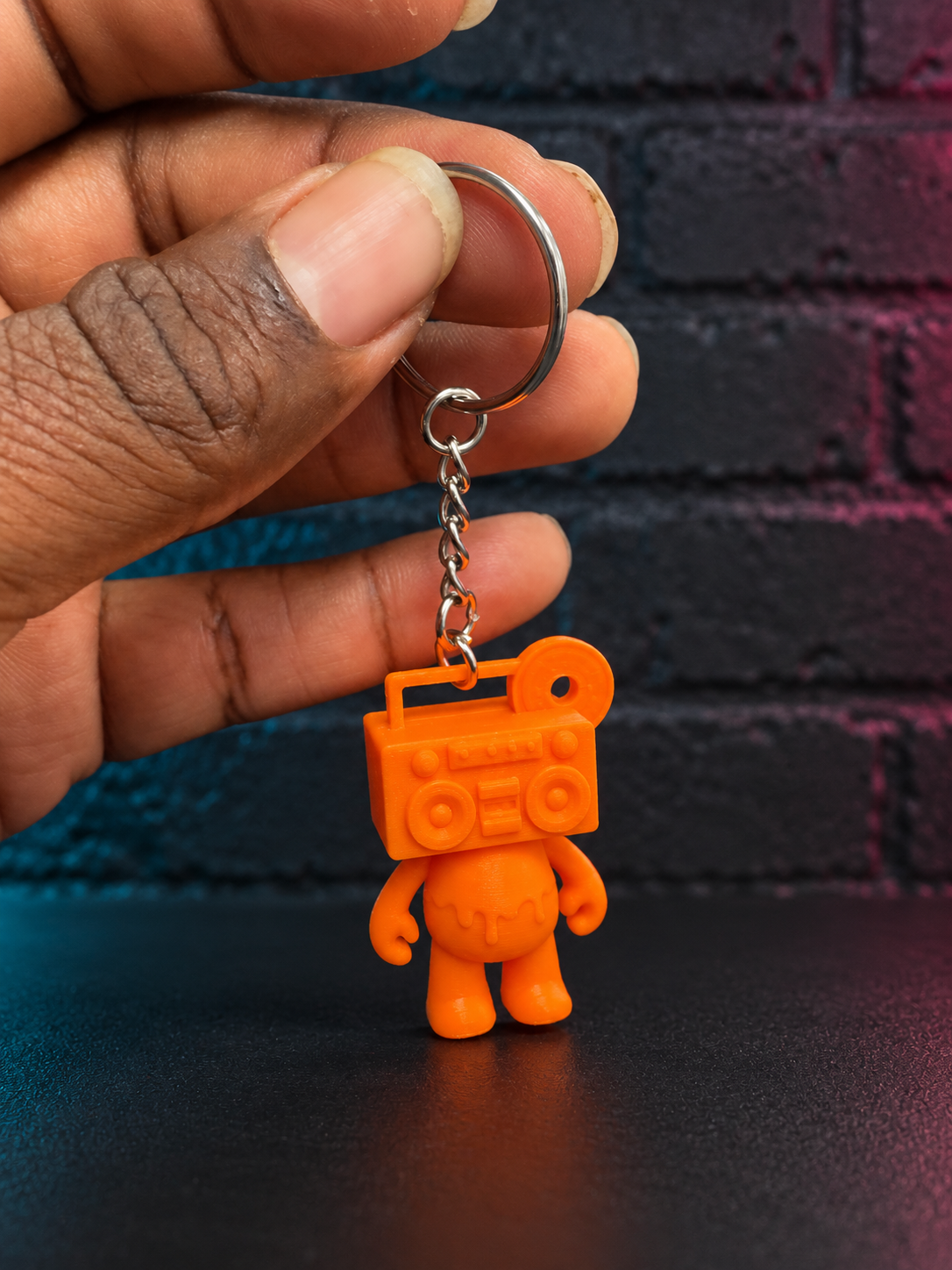 Electric Orange Boom Bear Mixtape Head keychain held by a Black hand against a charcoal brick wall with subtle cyan and magenta lighting