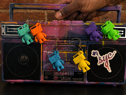 Black hand lifting a painted stereo displaying six colorful Boom Bear Mixtape Head keychains against a brick wall